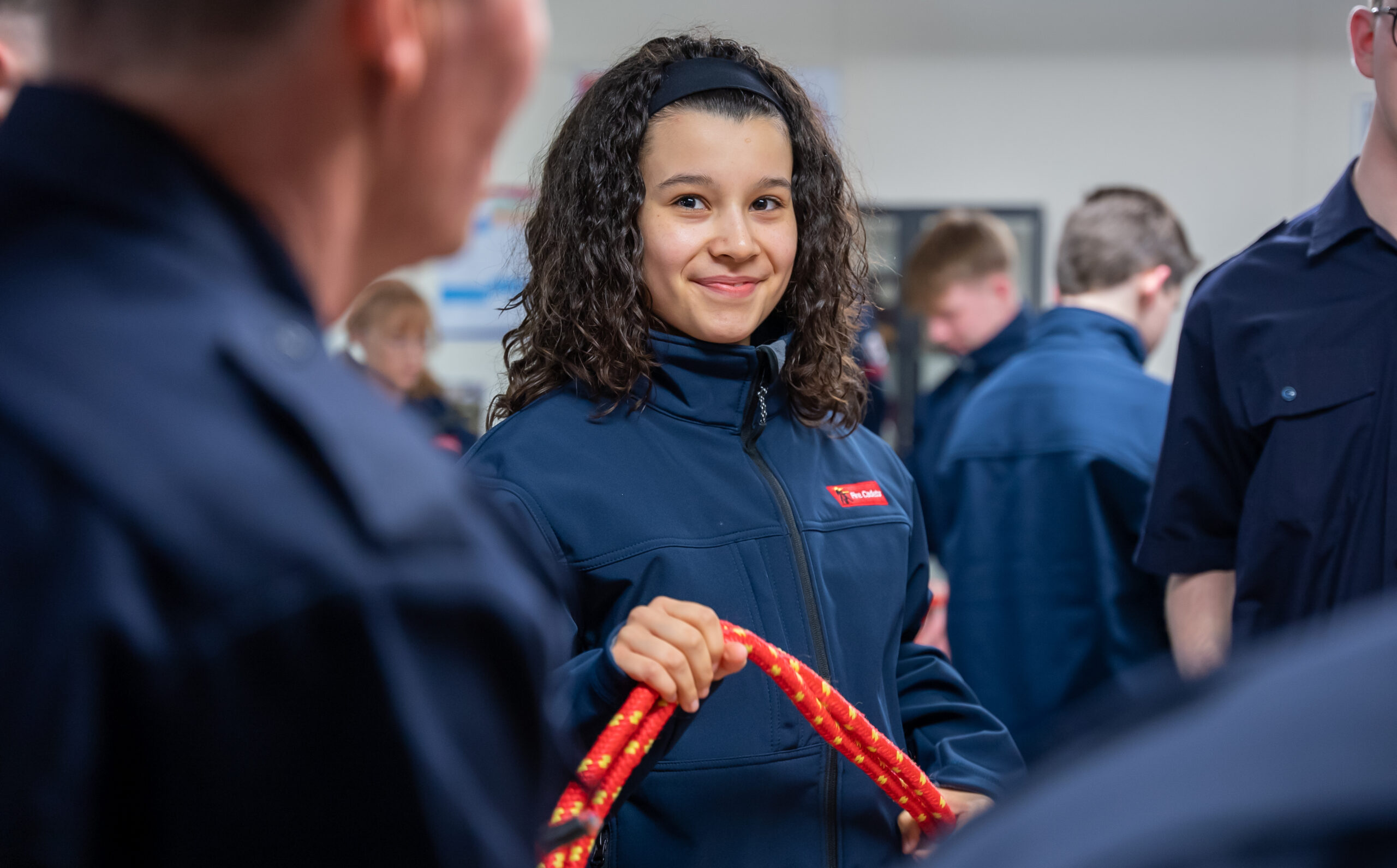 A Fire Cadet is smiling and looking at the camera while holding a length of rope. She is in a classroom with other Fire Cadets and they are all wearing blue uniforms.