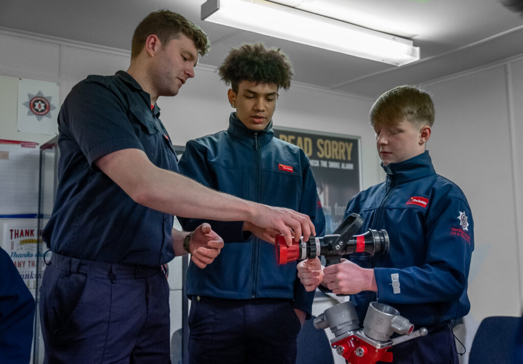 Photograph of a Fire Cadet Leader and two Fire Cadets wearing blue uniform in a classroom learning about a piece of equipment.