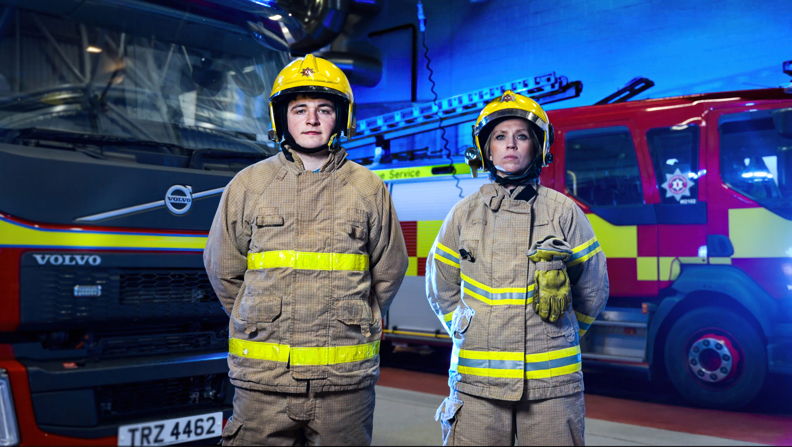 One male Firefighter and one female Firefighter standing in Firefighting uniform looking straight to camera with two fire appliances in the background.
